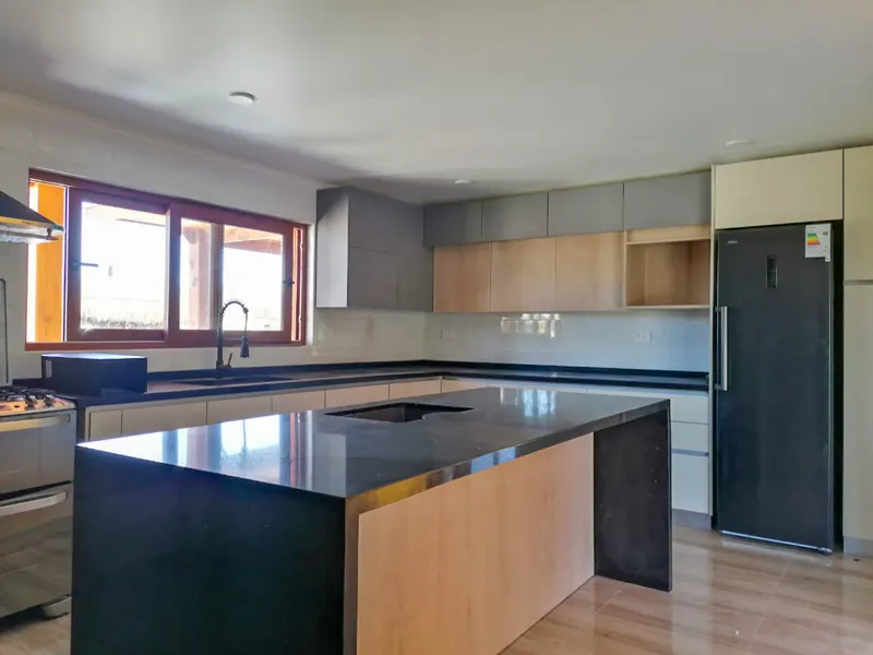 Cocina amplia con muebles bajos color arena, muebles altos grises y de madera clara, e isla central con cubierta de piedra oscura y lavaplatos integrado